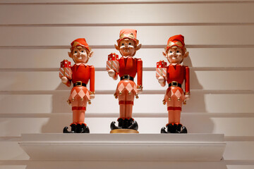 Three festive Christmas elf figurines holding gift boxes displayed on a white shelf. Holiday decoration concept with warm light, seasonal cheer, winter celebration, and traditional Christmas decor.