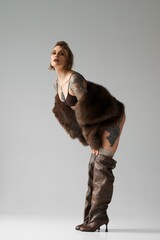 Woman with tattoos wearing a fur coat and thigh-high boots posing against a grey background