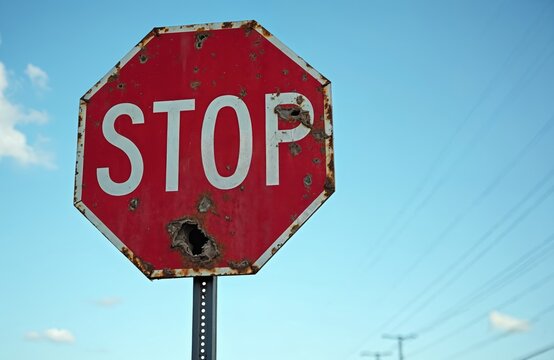 Damaged stop sign stands against blue sky. Rusty red roadsign is riddled with holes. Symbol of neglect and at city street warns drivers about safety.