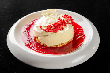 A close-up, high-angle studio shot of a gourmet white cheesecake with a cookie crust, topped with raspberry coulis and freeze-dried raspberries on a modern white plate