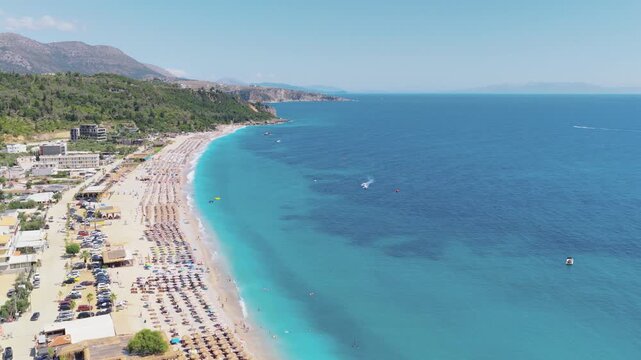 Aerial view of the vibrant Himare beach, dotted with umbrellas and people, contrasting against the deep blue sea, Himare, Vlore County, Albania.