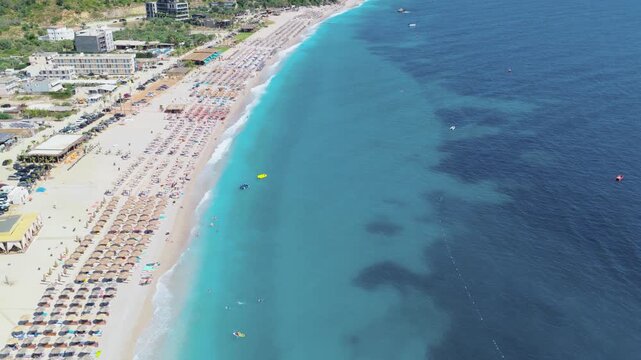 Aerial view of the turquoise sea meeting the sandy Himare beach packed with sunbathers and umbrellas, Himare, Vlore County, Albania.