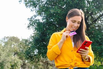 Happy woman wearing yellow shirt standing outdoors holding smartphone and credit card in green park, concept of online shopping, payment, banking, finance, e commerce, technology, freedom.Copy space