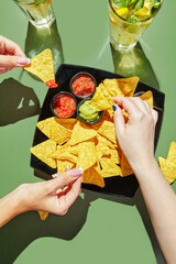 Female hands holding nachos corn chips and dipping them in salcha and guacamole sauces, glasses with lemonade are standing nearby. Mexican cuisine concept
