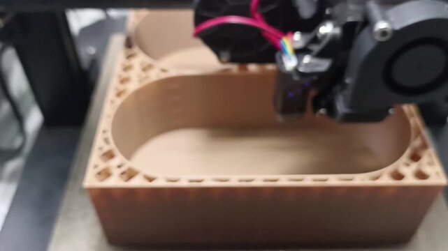 Detailed close-up shot of a Fused Deposition Modeling FDM 3D printer nozzle extruding light brown filament, building a complex container layer by layer.