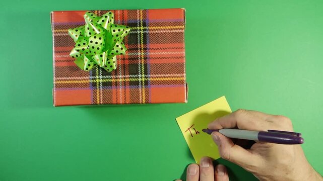 Writing "Thank You" Note Next to Plaid Gift Box; Top view of a hand writing a "Thank You" note on a yellow sticky paper next to a red plaid gift box with a green bow on a green screen background.