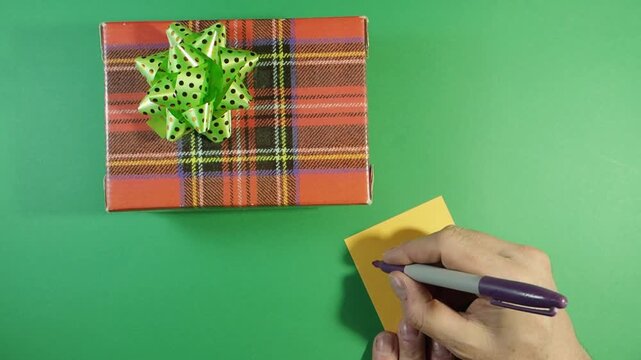 Writing "Thank You" Note Next to Plaid Gift Box; Top view of a hand writing a "Thank You" note on a yellow sticky paper next to a red plaid gift box with a green bow on a green screen background.