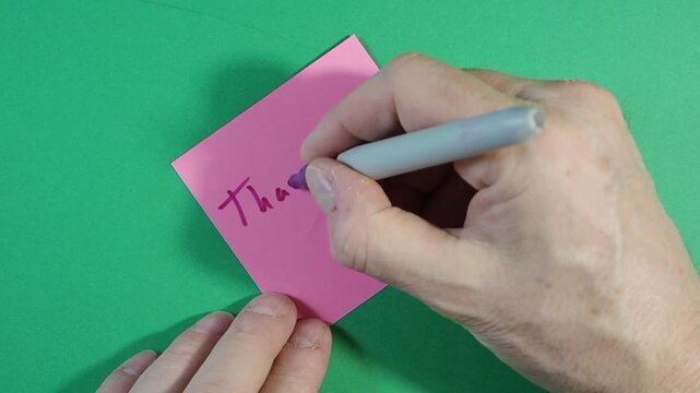 Writing "Thank You" on Pink Sticky Note; Close-up time-lapse of a hand writing the message "Thank You!" and drawing a smiley face on a pink sticky note with a marker, isolated on a green screen.