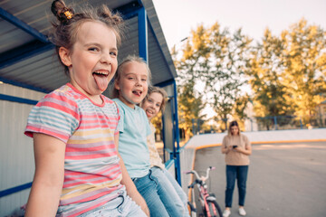 Three young girls sit on a bench, smiling and enjoying their time together. woman stands nearby, watching them. The scene captures family bonding and quality time.