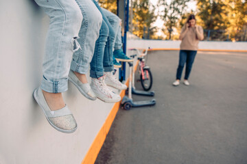 A group of children sitting on a wall in a court. They wear casual clothing and shoes. An adult woman stands nearby, talking on a phone. Trees are visible in the background.