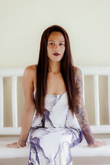 Tattooed woman seated with confident posture in printed dress, indoor editorial fashion portrait with soft tones
