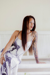 Elegant tattooed woman looking sideways while seated indoors, stylish fashion dress and soft minimalist space