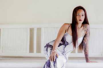 Stylish tattooed woman in long printed dress posing confidently in indoor setting with soft natural light