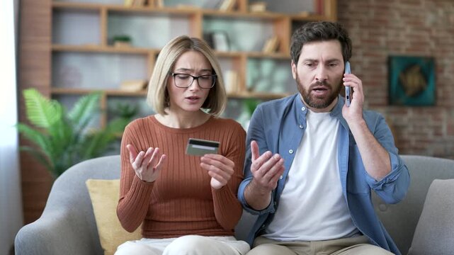 Confused married couple victim of credit card fraud sitting on sofa in living room at home. Disappointed angry husband and wife call the bank on phone and complain about problems and scam with payment