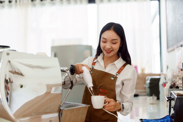 Young female barista making coffee, smiling while frothing milk in a white cup, working in a modern cafe