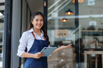 Young Asian woman, small business owner, standing outside coffee shop entrance, holding a tablet and gesturing welcome