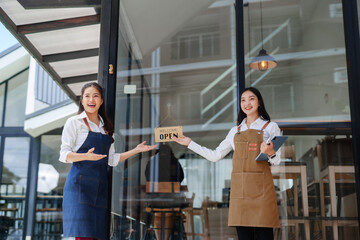 Two Asian women wearing aprons standing at their cafe entrance, holding an open sign and welcoming new clients