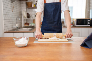 Man wearing apron rolling out dough on a wooden board at home, following a recipe from a tablet for a hobby cooking session