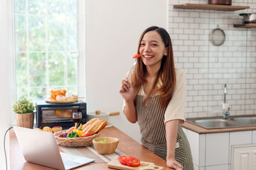 Young Asian woman smiling, holding a fork with a tomato slice in a bright kitchen while preparing a healthy meal