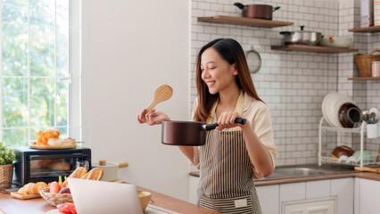 Young Asian woman cooking in kitchen, holding a saucepan and wooden spoon, learning a recipe while watching an online class