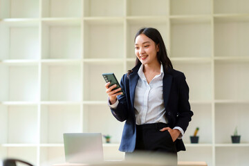 Young Asian businesswoman standing, smiling while using a smartphone, connected and communicating in a contemporary office