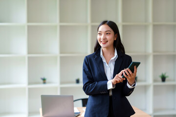 Asian businesswoman in a suit smiling, holding a smartphone, and looking away in a bright, modern office environment