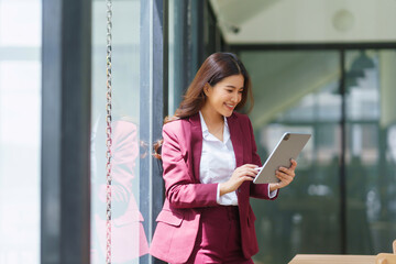 Young Asian businesswoman in a suit smiling, working with a tablet, standing in a contemporary office setting