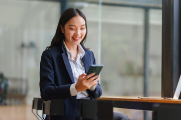 Asian businesswoman smiling, interacting with a smartphone in a modern office, representing communication and digital connectivity