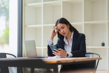 Young professional woman feeling overwhelmed and frustrated, holding her phone with a headache while working in a modern office