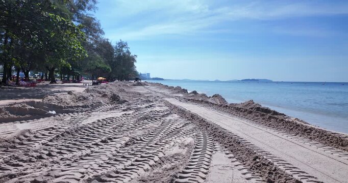 Shoreline construction scene featuring heavy tread marks and distant excavator against a calm blue sea.