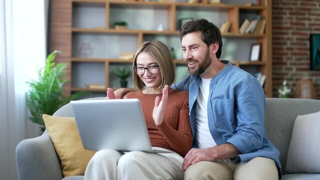 Satisfied happy married couple received great news on laptop while sitting on sofa in living room at home. Smiling glad husband and wife reads a positive good message looking at the computer screen