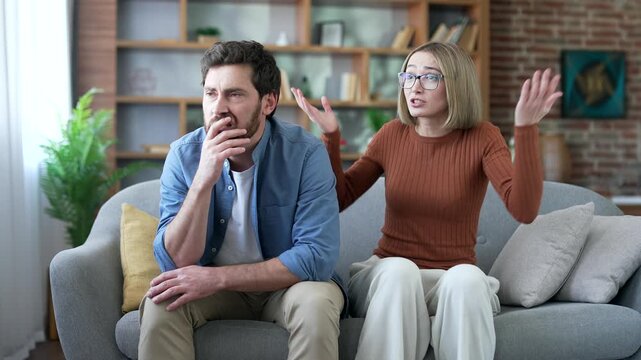 Angry wife arguing with sad husband sitting on sofa in living room at home. Conflict and dispute between a married couple. A woman quarrels with a depressed man. Problem and crisis in family relations