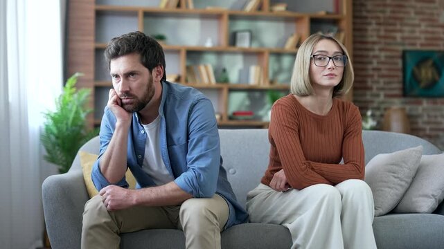 Upset quarreling wife and husband, offended at each other, sitting side by side on sofa at home. Depressed man and woman are angry. Conflict in a married couple. Problem, crisis in family relations