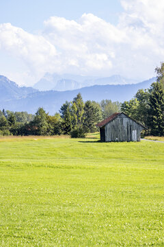 View of a weathered wooden shack with a red-tinged roof sits amid a vibrant green field under a bright sky, with distant, hazy mountains, Zug, Switzerland.