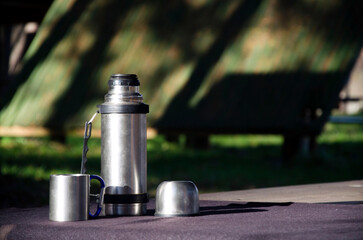 Stainless steel thermos with an open lid and metal cup placed on a table outdoors in natural light.