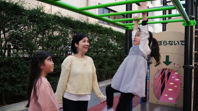 Young Asian girl playing on monkey bars with her mother and sister encouraging her in a city park