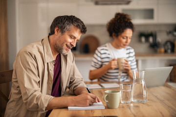 Couple At Kitchen Table Working Together: Man Writing Notes, Woman Drinking Coffee