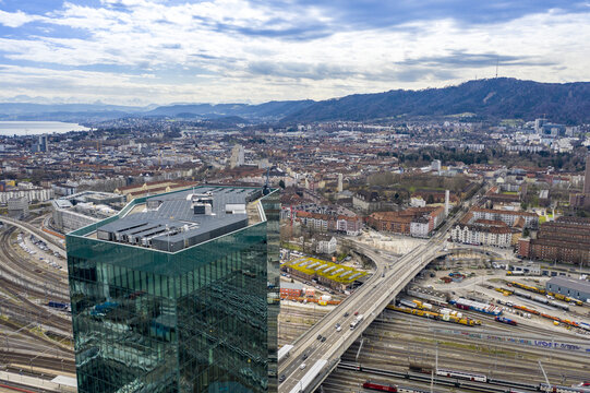 Aerial view of a modern glass skyscraper contrasting with the sprawling cityscape and distant mountains under a textured sky, Flawil, St. Gallen, Switzerland.