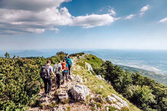 View of hikers trek along a narrow, rocky path atop a mountain ridge, vibrant green foliage contrasting with the clear blue sky, Mala Ucka, Primorje-Gorski Kotar County, Croatia.