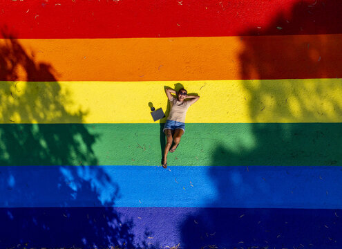 Aerial view of a person lying on a vibrant rainbow-colored pride path, bathed in sunlight and dappled with shadows, creating a striking contrast of light and color, Melbourne, Victoria, Australia.
