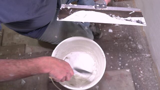A professional worker uses a taping knife to prepare drywall mud or spackle, scooping the material from a bucket for wall finishing.