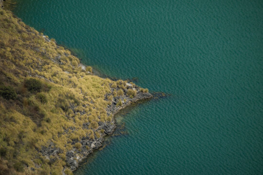 View of the turquoise lake water meeting the rugged, golden vegetated shoreline, creating a striking contrast of colors and textures, Quilotoa lake, Cotopaxi province, Ecuador.