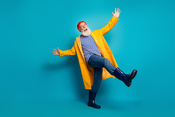 Happy senior man in yellow raincoat dances with striped shirt jeans and beanie in a bold studio setting against a bright blue background
