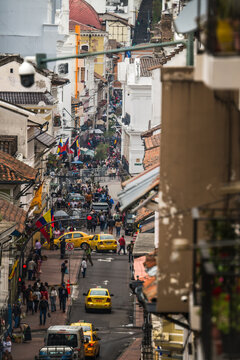 View of a bustling street descends through weathered buildings, colorful flags flutter amidst a vibrant tapestry of vehicles and pedestrians, Quito, Ecuador.