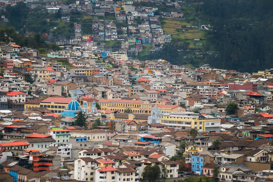 View of a densely packed cityscape of terracotta roofs and colorful buildings, nestled against verdant hillsides under a vast sky, Quito, Ecuador.