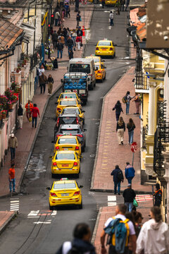 View of a vibrant street scene unfolds with yellow taxis lining the cobblestone road, framed by colorful buildings and bustling with people, Quito, Ecuador.