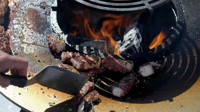 Close-up of a person using a metal spatula scraper to move pieces of octopus or meat around the hot, seasoned surface of a circular fire pit grill over open flames for even cooking.