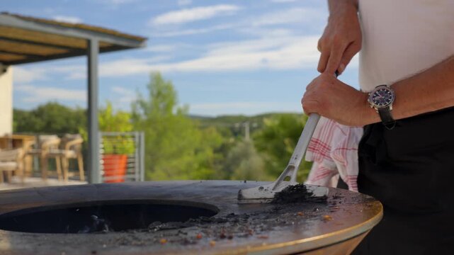 Person cleaning a round outdoor grill by pushing leftover ash and food residue toward the central fire pit using a metal scraper. Barbecue maintenance during outdoor cooking in a backyard setting.