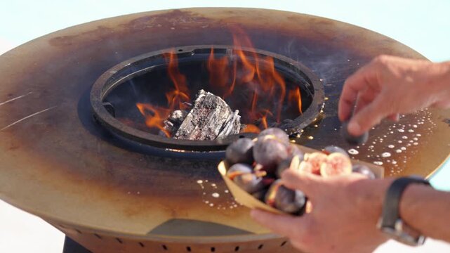 A person's hands place fresh figs onto the hot, circular surface of an outdoor fire pit grill with an open fire burning in the center. Focus on gourmet grilling and cooking outdoors.
