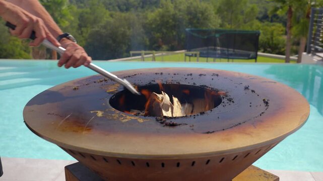 Close-up of a person tending a modern barbecue fire pit with metal tongs, beside a swimming pool. Summer leisure, outdoor cooking, relaxation, and backyard lifestyle.
